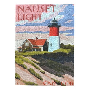 May include: A completed jigsaw puzzle featuring the Nauset Light lighthouse on Cape Cod. The lighthouse is red and white, with a black top. A house with a red roof sits nearby. The sky is blue and pink.