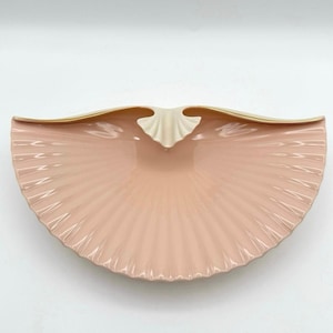 May include: A semi-circular ceramic dish shaped like a fan or a seashell, featuring a soft pastel pink color with a glossy finish. The top edge is accented with a thin, smooth gold-toned trim, and the center displays a small, cream-colored detail.