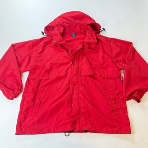 May include: A bright red hooded jacket with a snap and zipper closure. The jacket has two front pockets and a drawstring at the hood and hem. The jacket is made of a lightweight material.