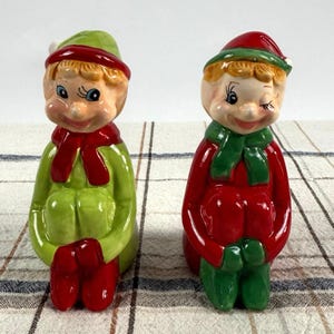 Op de afbeelding: Twee keramische elfenbeeldjes. De ene elf draagt een groene outfit met een rode sjaal en hoed, de andere draagt een rode outfit met een groene sjaal en hoed. Beide hebben roze wangen en lachen.