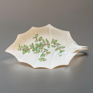 May include: A cream-coloured, leaf-shaped serving dish with a handle. The dish is decorated with a green holly and red berry design. The rim of the dish has a gold trim. A festive piece for serving snacks or desserts.