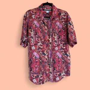 Peut inclure: Une chemise à manches courtes avec un motif audacieux en rouge, violet et beige. La chemise est suspendue à un cintre noir. Le motif présente des formes géométriques et abstraites. L'arrière-plan est de couleur pêche.