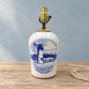 May include: A white ceramic table lamp base featuring a blue ink-style illustration of a lighthouse and buildings. The text CHATHAM LIGHT CAPE COD is printed below the image. The lamp has a brass-colored metal socket and a white electrical cord.