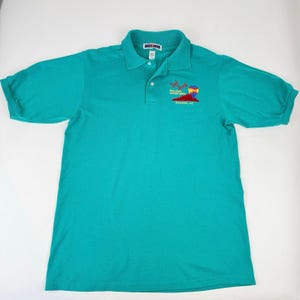 May include: Teal polo shirt with short sleeves and a collared neckline. Features a small embroidered design of a hot air balloon and text that reads "Red Rock Balloon Adventures, Sedona, AZ". The shirt has a three-button placket.