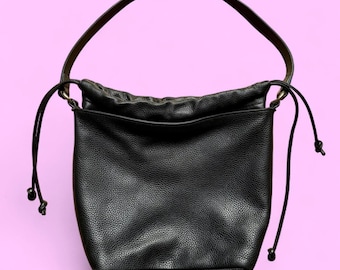 May include: A black leather bucket bag with a drawstring closure and a single shoulder strap. The bag has a textured surface and a simple, elegant design. The background is a solid light purple.