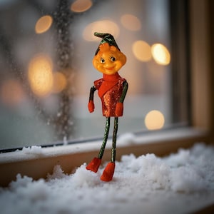 Puede incluir: Una caprichosa figura de elfo navideño con una cara sonriente, vestida con un atuendo rojo con lentejuelas, piernas verdes y negras y botas rojas. El elfo tiene un sombrero verde y naranja. La figura está sobre un alféizar nevado.