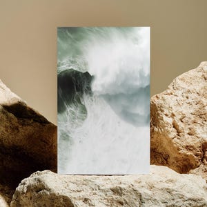Puede incluir: Una impresión artística rectangular que muestra una dramática ola oceánica en tonos verdes y blancos. La impresión se exhibe sobre un fondo beige, sostenida por rocas grandes y texturizadas.