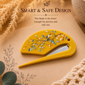 May include: A yellow semi-circular safety cutter featuring a delicate blue and orange leaf pattern on its surface. The tool includes a small hole for hanging and a sharp metal blade positioned in the lower triangle for precise cutting.