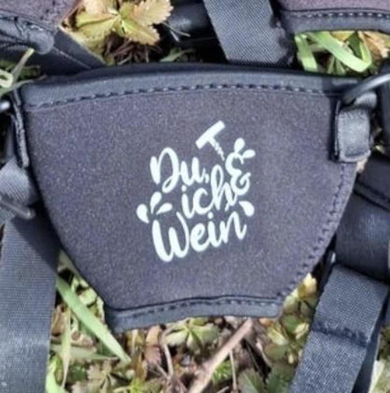 Ein schwarzer Weinglashalter mit dem deutschen Text "Du, ich & Wein" in weißer Schrift. Der Träger ist von schwarzen Riemen umgeben und liegt auf einem Bett aus grünem Gras und Blättern.