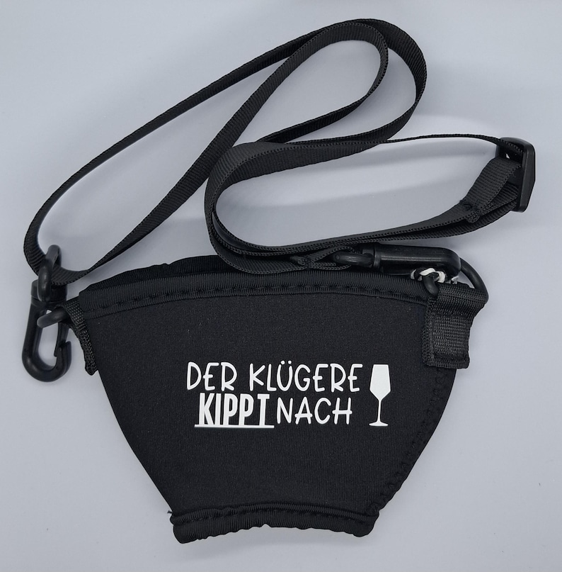 Schwarzer Neopren-Weinglashalter mit schwarzem Riemen und Clip. Der Halter hat weißen Text "DER KLÜGERE KIPPI NACH" und eine Weinglas-Grafik. Der Riemen ist verstellbar und kann um den Hals getragen werden.