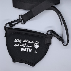 Schwarzer Weinglashalter mit schwarzem Gurt und Clips. Der Halter hat weißen Text mit der Aufschrift "DIE tut nix, die will nur WEIN" und eine Weinglasgrafik mit Herzen.