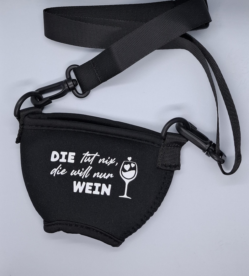Schwarzer Weinglashalter mit schwarzem Gurt und Clips. Der Halter hat weißen Text mit der Aufschrift "DIE tut nix, die will nur WEIN" und eine Weinglasgrafik mit Herzen.