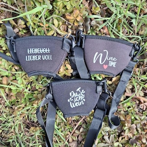 Drei schwarze Weinglashalter mit verstellbaren Trägern werden auf einem Bett aus Gras und Blättern gezeigt. Jeder Halter hat weißen Text: "LIEBEVOLL LIEBER VOLL", "Wine Time" mit einem Herz und "Du & ich & Wein".