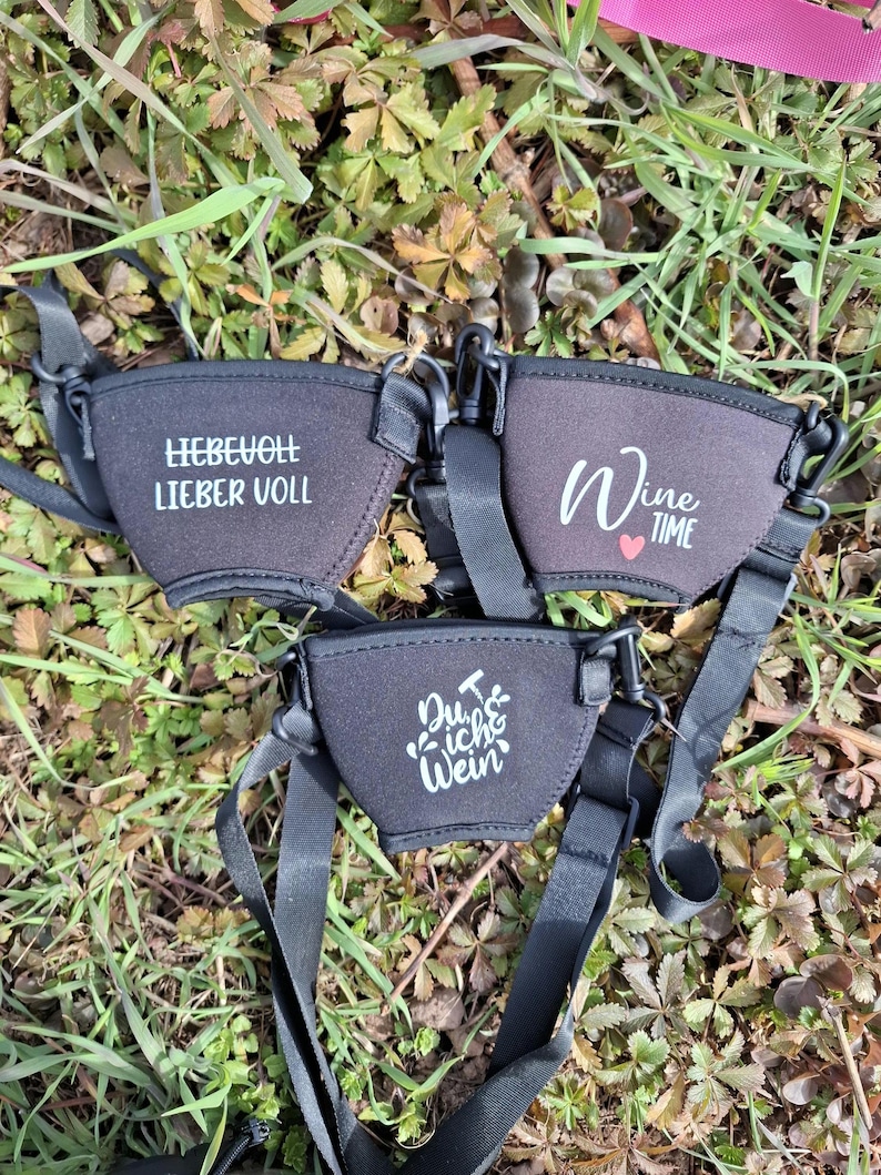 Drei schwarze Weinglashalter mit verstellbaren Trägern werden auf einem Bett aus Gras und Blättern gezeigt. Jeder Halter hat weißen Text: "LIEBEVOLL LIEBER VOLL", "Wine Time" mit einem Herz und "Du & ich & Wein".