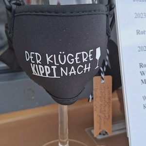 Ein Weinglas mit einer schwarzen Stoffhülle mit dem weißen Text "DER KLÜGERE KIPPT NACH".