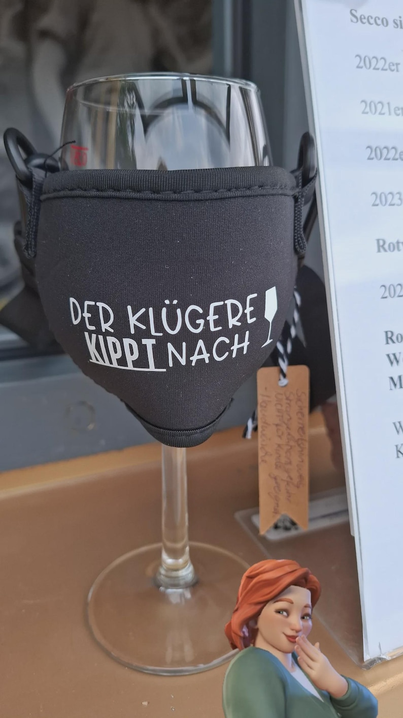 Ein Weinglas mit einer schwarzen Stoffhülle mit dem weißen Text "DER KLÜGERE KIPPT NACH".