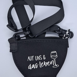 Schwarzer Weinglashalter mit schwarzem Riemen und Verschluss. Der Halter hat den Text "AUF UNS & das leben" und eine Weinglasgrafik in Weiß. Der Riemen ist verstellbar.