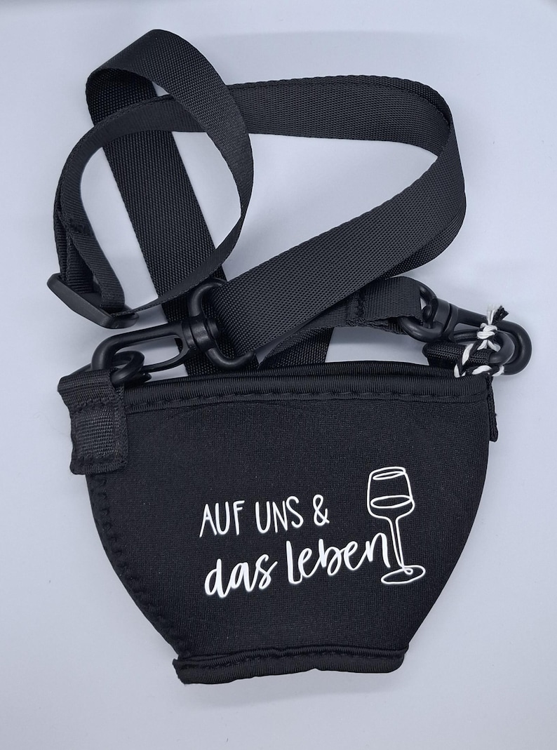 Schwarzer Weinglashalter mit schwarzem Riemen und Verschluss. Der Halter hat den Text "AUF UNS & das leben" und eine Weinglasgrafik in Weiß. Der Riemen ist verstellbar.