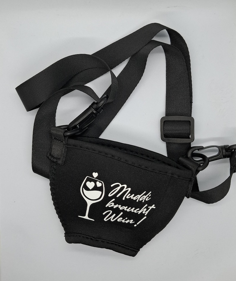 Schwarzer Weinglashalter mit einem schwarzen, verstellbaren Riemen. Der Halter hat den Text "Muddi braucht Wein!" und eine Weinglasgrafik mit Herzen. Der Halter ist für das Tragen eines Weinglases konzipiert.