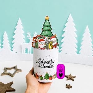 Könnte beinhalten: Ein zylindrischer Adventskalender aus Papier mit der Aufschrift Advents kalender in schwarzer Schreibschrift. Das Design zeigt bunte Illustrationen eines Weihnachtsbaums, des Weihnachtsmanns und eines Pinguins sowie die Zahlen 5, 4 und 6.