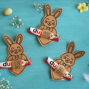 Può includere: Decorazioni pasquali con coniglietti di carta marrone che tengono barrette di cioccolato rosse e bianche. I coniglietti hanno corpi a forma di cuore e papillon. Il testo include "Frohe Ostern", "Oster Hasi" e "Bist Süss".