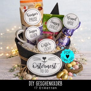 Może przedstawiać: Ciemnoszary filcowy kosz wypełniony wielkanocnymi smakołykami i oznaczonymi przedmiotami. Kosz zawiera kubek, słodycze i czekoladowego zajączka. Etykiety zawierają frazy takie jak "Osternest to Go" i "Easter Light".