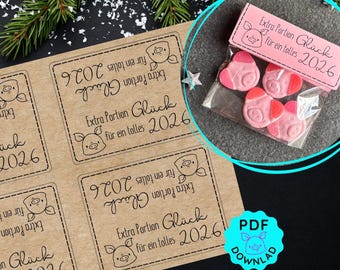 May include: A festive image featuring a clear bag filled with pink pig-shaped candies, labeled "Extra Portion Glück für ein tolles 2026." Also shown are kraft paper labels with the same text and pig illustration, and a PDF download icon.