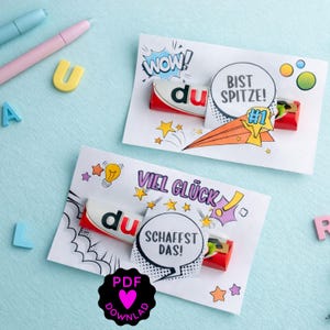 Può includere: Due biglietti bianchi con testo tedesco e barrette di cioccolato. Un biglietto dice "WOW!" e "BIST SPITZE! #1". L'altro dice "VIEL GLÜCK" e "SCHAFFST DAS!". Un'icona di download PDF nera è in basso a sinistra.