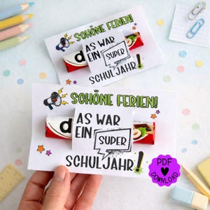 Pode incluir: Dois cartões brancos com o texto "SCHÖNE FERIEN!" e "AS WAR EIN SUPER SCHULJAHR!". Cada cartão tem uma barra de chocolate anexada. Os cartões são decorados com estrelas e gráficos de megafone. Lápis e confetes no fundo.
