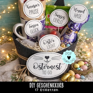Peut inclure: Un panier en feutre gris rempli de friandises de Pâques, dont une tasse violette, un lapin en chocolat et des bonbons. Le panier comporte des étiquettes décoratives avec du texte allemand comme "Osternest TO GO" et "Knusper PAUSE". Un petit nid d'œufs colorés est également inclus.