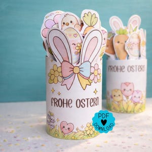 Op de afbeelding: Twee witte cilindrische containers versierd met paasillustraties. Eén heeft konijnenoren, een strik en de tekst "FROHE OSTERN". De andere heeft wortel- en ei-ontwerpen. Beide zijn gevuld met paasuitsparingen. Een PDF-downloadpictogram is zichtbaar.