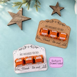 Puede incluir: Dos tarjetas decorativas con el texto "DREI HASELNÜSSE FÜR DICH" y "Wünsch Dir was!" con tres bombones Ferrero Küsschen pegados en cada una. Las tarjetas van acompañadas de dos adornos de estrellas de madera sobre un fondo azul claro.