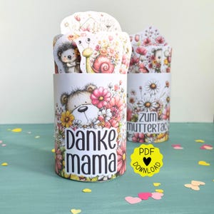 May include: Two cylindrical gift boxes with floral and animal illustrations. One box says "Danke Mama" and the other says "Zum Muttertag". The boxes are decorated with pink, yellow, and white flowers, and feature a teddy bear, hedgehog, and snail.