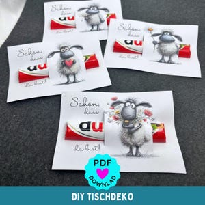 May include: Several handmade greeting cards with illustrations of sheep holding flowers, hearts, and daisies. Each card has a Kinder Bueno chocolate bar attached. The cards have German text and a turquoise PDF download graphic.