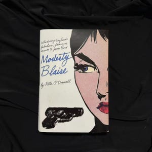 Puede incluir: Un libro de bolsillo vintage titulado "Modesty Blaise" de Peter O'Donnell. La portada presenta una ilustración de estilo pop art del rostro de una mujer con labios rojos y ojos azules, y una pistola negra. El título del libro está en escritura azul.