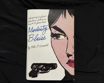 May include: A vintage paperback book titled "Modesty Blaise" by Peter O'Donnell. The cover features a pop art style illustration of a woman's face with red lips and blue eyes, and a black handgun. The book's title is in blue script.