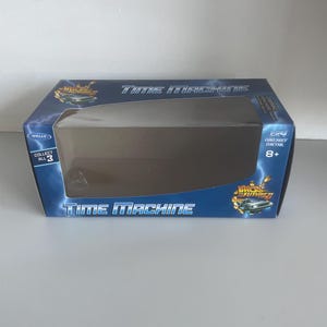 May include: A blue box with the words "Time Machine" in white. The box has a clear plastic window and features the "Back to the Future" logo. The box also says "Collect All 3" and "8+".