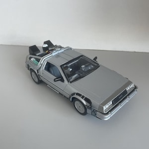 May include: Silver DeLorean model car with black accents, featuring detailed headlights, wheels, and a distinctive rear section. The model car is a replica of the iconic vehicle from the Back to the Future film series.