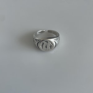 Peut inclure: Une bague argentée avec un design ouvert. La bague présente un emblème central avec un design stylisé, et la bande est gravée de "S925". La bague est posée sur un fond uni.