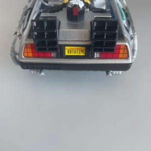 May include: A silver DeLorean model car with black vents and red and orange tail lights. The license plate reads "OUTATIME" in yellow. The car has a detailed engine compartment and is set against a light gray background.