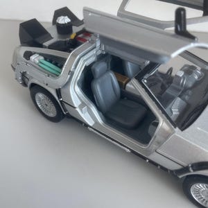May include: A silver die-cast model of a DeLorean car with gull-wing doors open. The interior features gray seats and detailed dashboard. The rear compartment is visible, with various components and tubes. The car's wheels have intricate spoke designs.