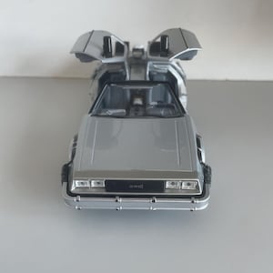 May include: Silver die-cast model car with gull-wing doors open. The car has a detailed interior and exterior, including headlights, taillights, and the DMC logo on the front. The car is a replica of the DeLorean.