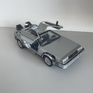 Puede incluir: Un modelo plateado de un coche DeLorean DMC-12 con las puertas de ala de gaviota abiertas. El coche en miniatura tiene detalles como ruedas, faros e interior. El modelo está sobre una superficie blanca.