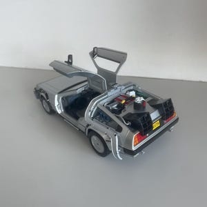 May include: A silver DeLorean model car with gull-wing doors open. The model has detailed features, including the engine and interior. The car has black and red taillights and a yellow license plate.