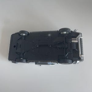 May include: A die-cast model car, viewed from the underside, showcasing intricate details. The car is primarily black with silver accents, featuring four visible wheels and detailed chassis components. The model sits on a light gray surface.