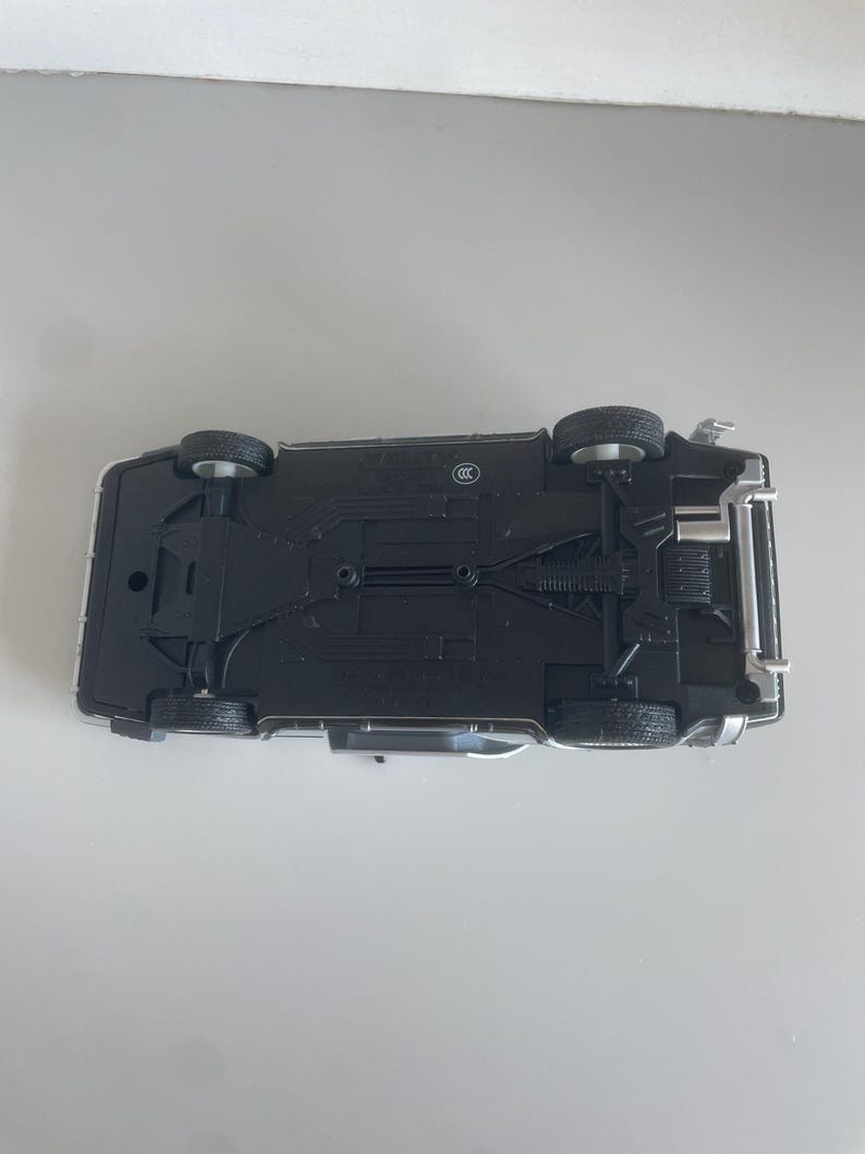 May include: A die-cast model car, viewed from the underside, showcasing intricate details. The car is primarily black with silver accents, featuring four visible wheels and detailed chassis components. The model sits on a light gray surface.
