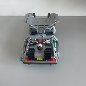 May include: A silver DeLorean model car with gull-wing doors open. The car has a detailed interior, including a red engine and the license plate reads "OUTATIME". The car is a replica of the vehicle from the Back to the Future movie.