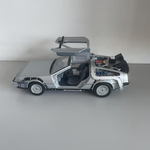 May include: A silver die-cast model of a DeLorean DMC-12 car with gull-wing doors open. The model features detailed interior and exterior components, including the flux capacitor and rear engine details. The car is approximately 10 inches long.