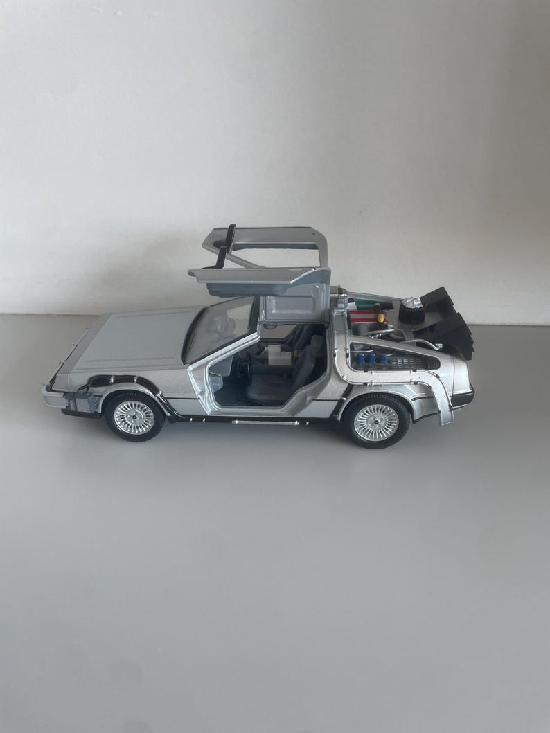 May include: A silver die-cast model of a DeLorean DMC-12 car with gull-wing doors open. The model features detailed interior and exterior components, including the flux capacitor and rear engine details. The car is approximately 10 inches long.