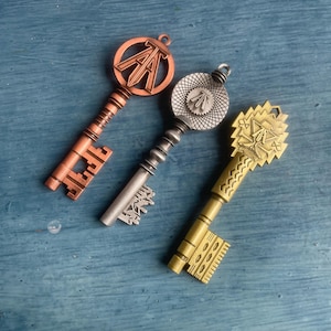 Op de afbeelding: Drie decoratieve metalen sleutels in koper, zilver en goudtinten. Elke sleutel heeft een uniek ontwerp op de kop en ingewikkelde details op de tanden. De koperen sleutel heeft de letters "A" en "T".